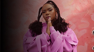 Singer Yemi Alade poses with her hands in a prayer formation in front of a pink wall